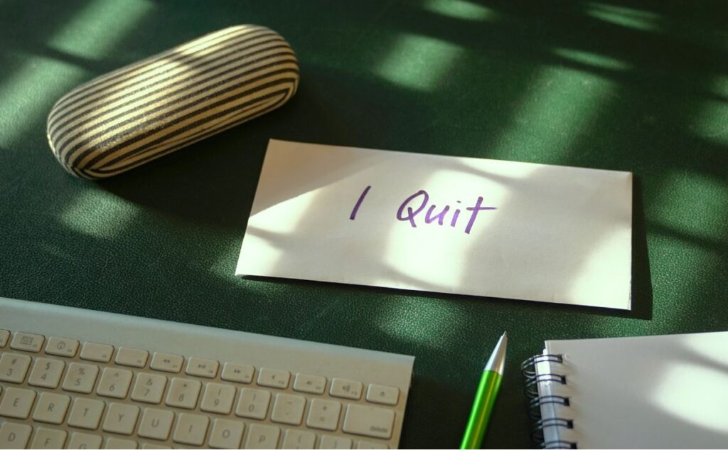 A handwritten note saying "I quit" lying on a green surface surrounded by desk equipment.