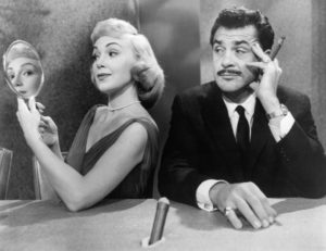 A woman turns away from an exasperated man with a mustache and cigar and looks at him through a hand held mirror